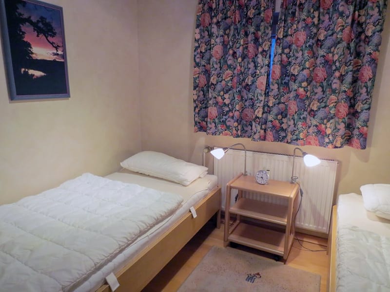 Schlafzimmer mit Einzelbett, Nachttischwagen mit Wecker, zwei Leselampen und zweitem Bett am rechten Bildrand