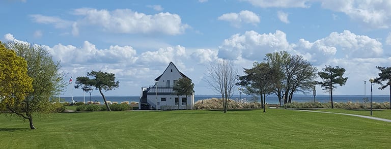 Rasenfläche mit einzelnen Bäumen, in der Mitte ein weißes Haus, dahinter ein Streifen Ostsee