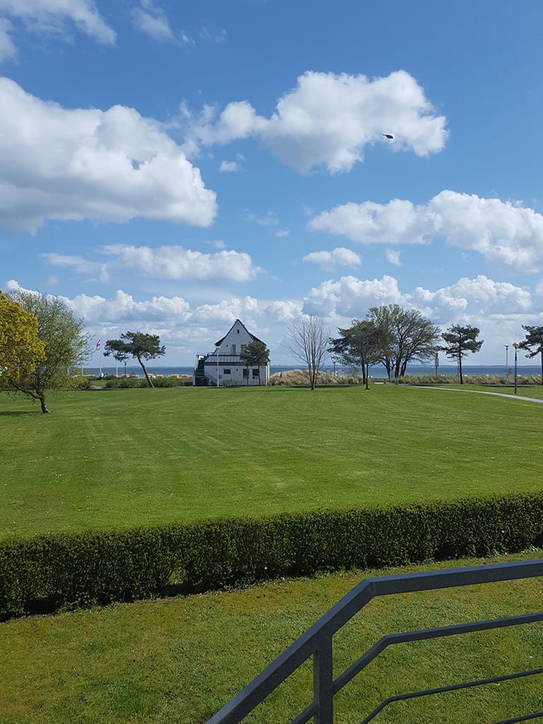 Rasenfläche mit einzelnen Bäumen, in der Mitte ein weißes Haus, dahinter ein Streifen Ostsee