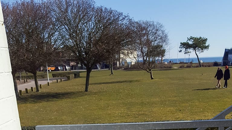 Sitzplatz im Freien mit Klapptisch und Gartenstuhl, dahinter Rasenfläche, Bäume und ein Streifen Ostsee am Horizont