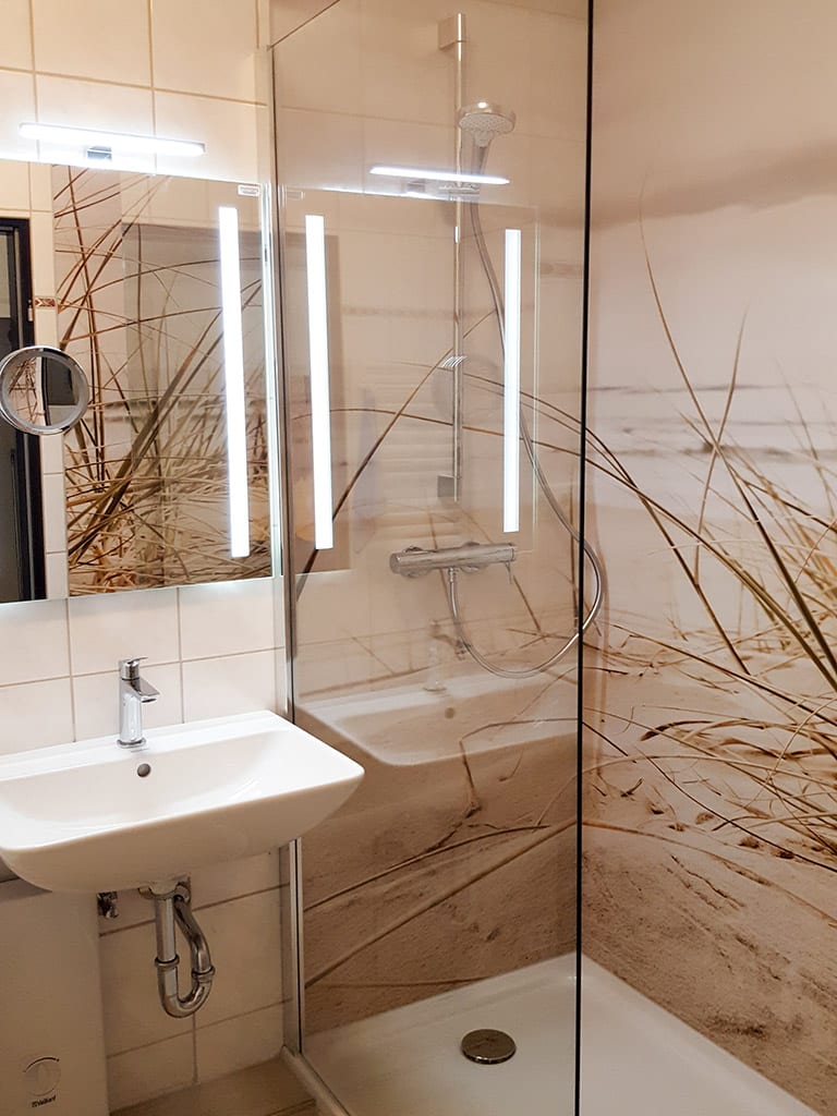 Duschbad mit Glasabtrennung, Dünenmotiv als Rückwand, Waschbecken und Spiegelleuchten